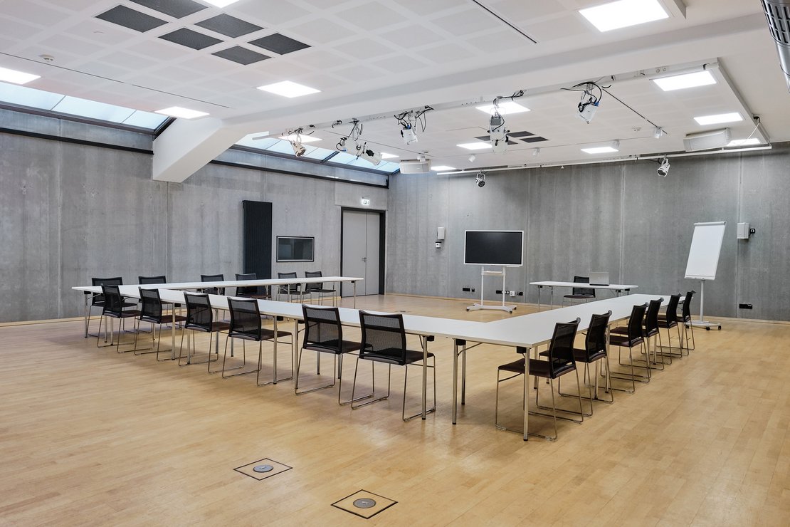 Ein modern eingerichteter Besprechungsraum mit schwarzen Stühlen an weißen Tischen als U aufgebaut und technischer Ausstattung an der Decke.
