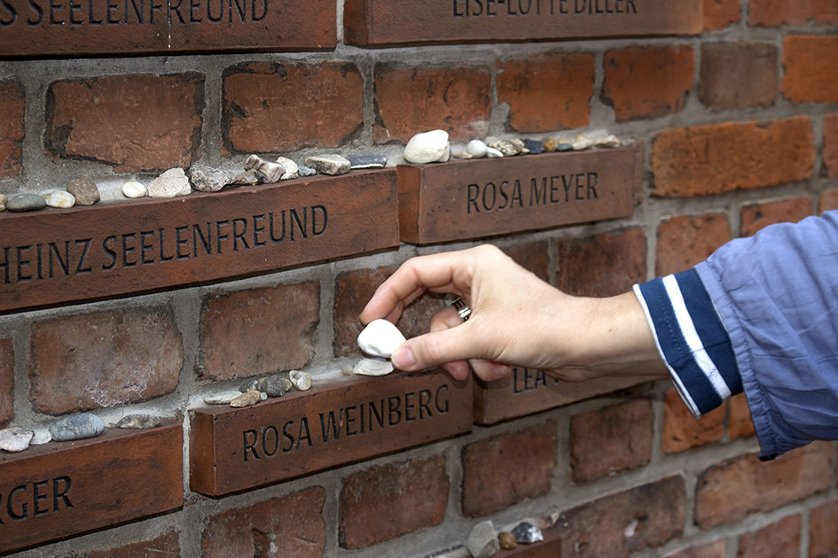 Aus der Gebäudemauer hervortretende Steine zeigen die Namen Rendsburger Juden. Kleine Gedenksteine werden darauf abgelegt.